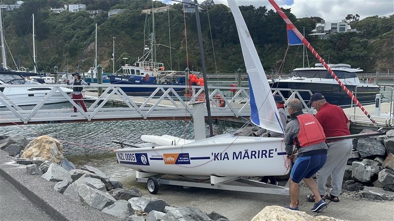 Two individuals moving a yacht by the water, with a bridge in front and a person standing on it